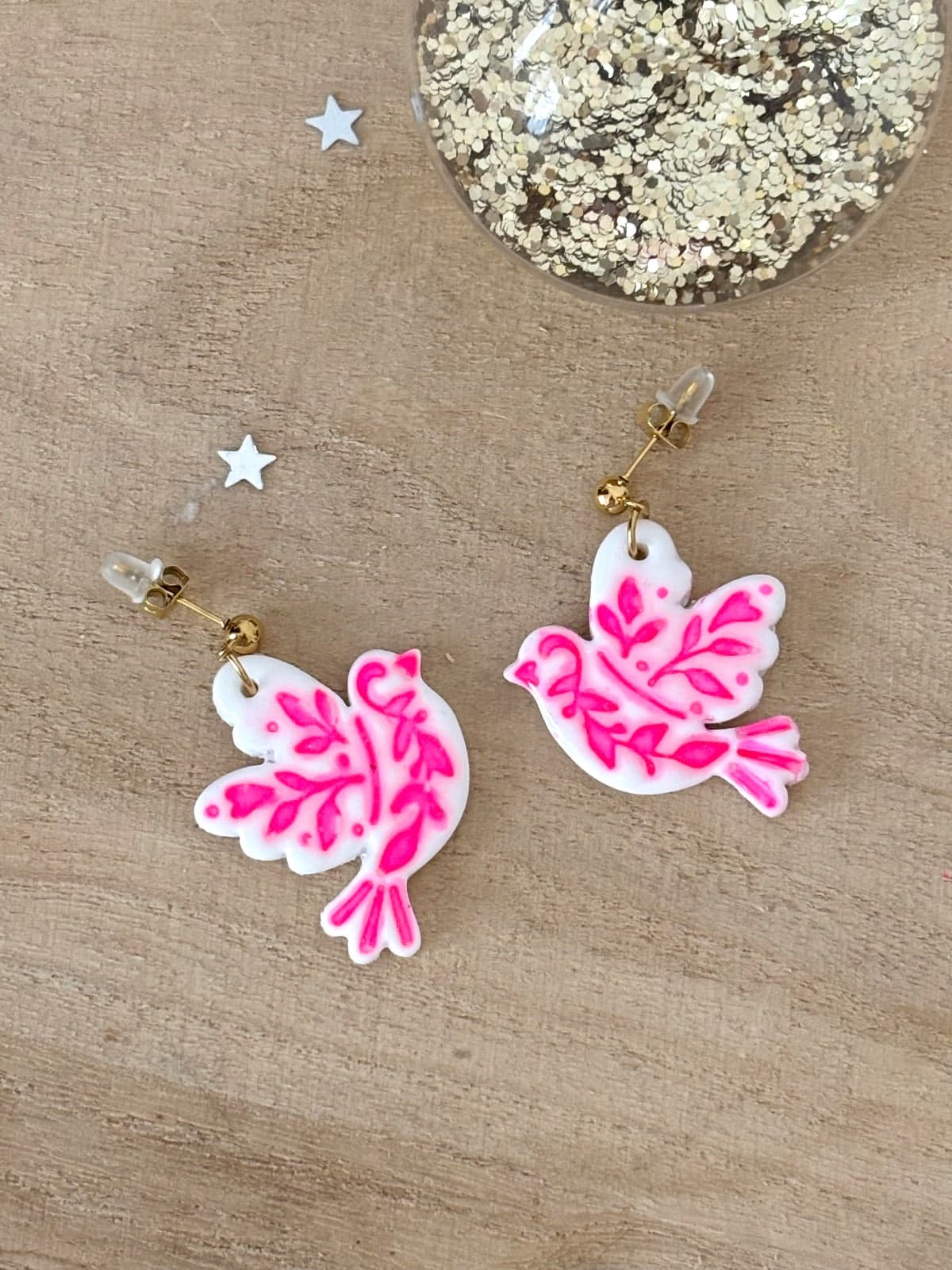 Paire de boucles d'oreilles oiseaux blancs à motifs rose vif posée sur fond en bois.